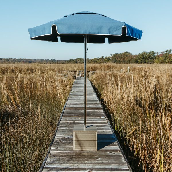 A Box Anchor Outdoor Umbrella Base sits on a dock with a Capri Palmetto Umbrella, marshland is seen behind it. 